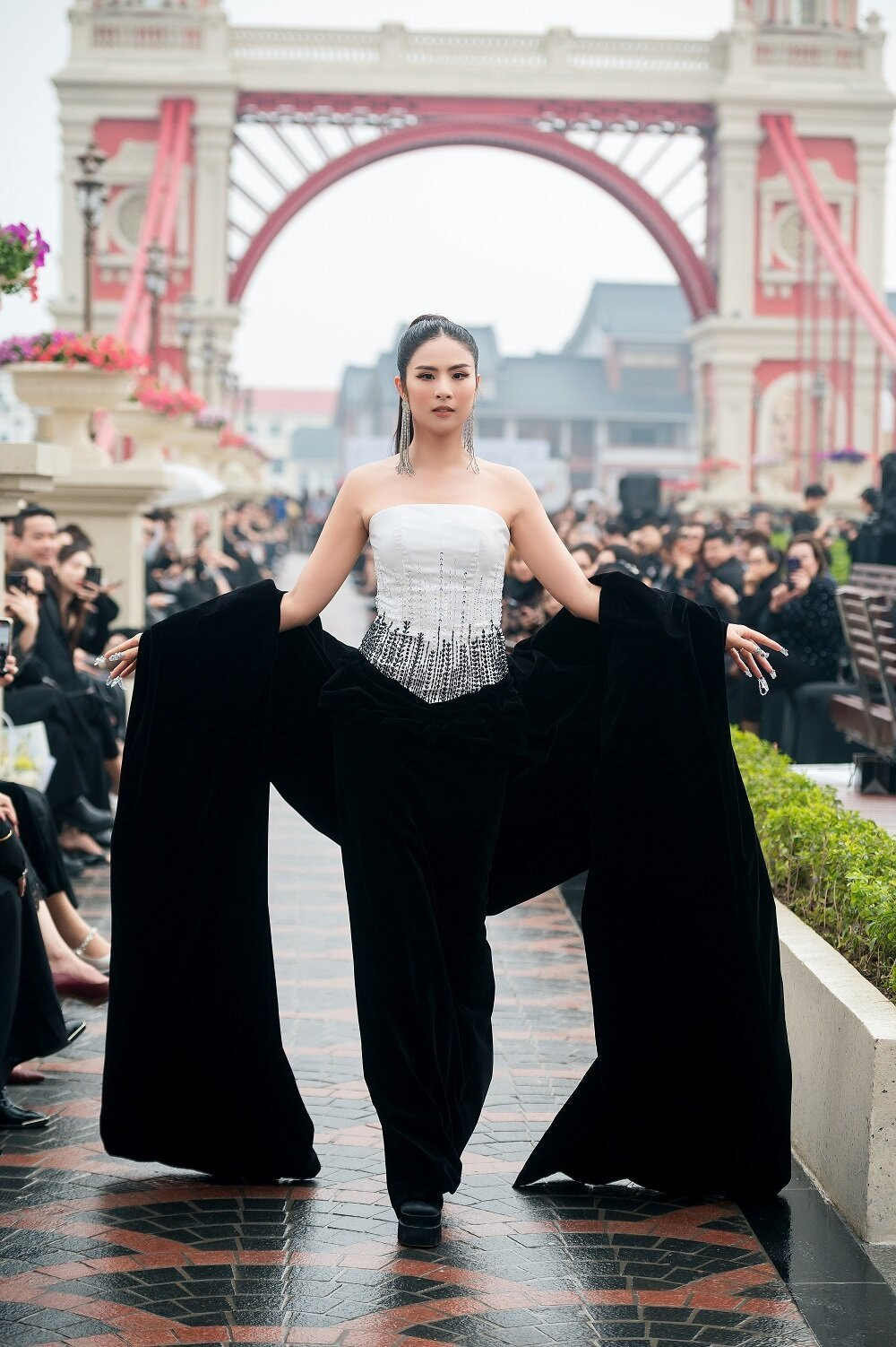Hoa hậu Việt Nam 2010 biểu cảm sắc lạnh, visual đầy cuốn hút, kiêu sa với kiểu tóc buộc đuôi ngựa hiện đại. Người đẹp sải bước trong bộ đầm tông đen trắng, quây ngực, đính cườm và sequin ở thân trên, nhấn bằng áo choàng quá khổ.